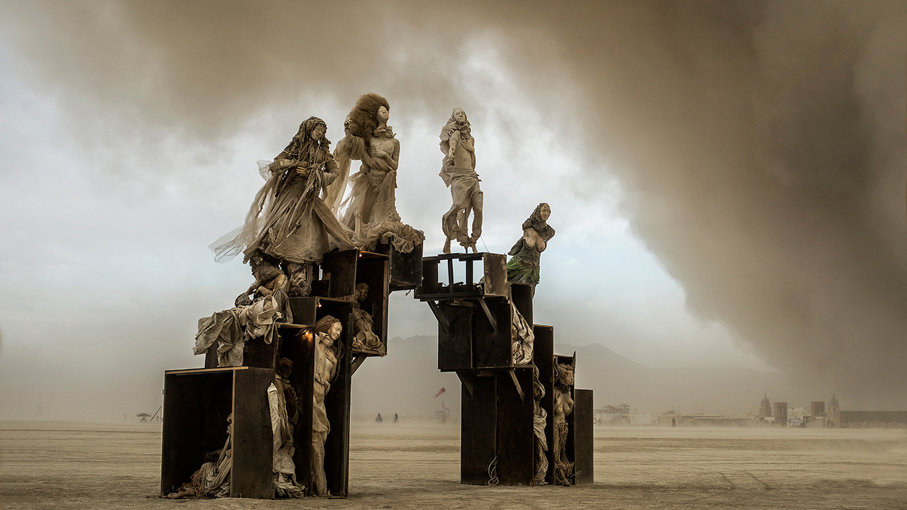 Robed figures perched atop weathered wooden crates on the playa, beneath an approaching dust storm