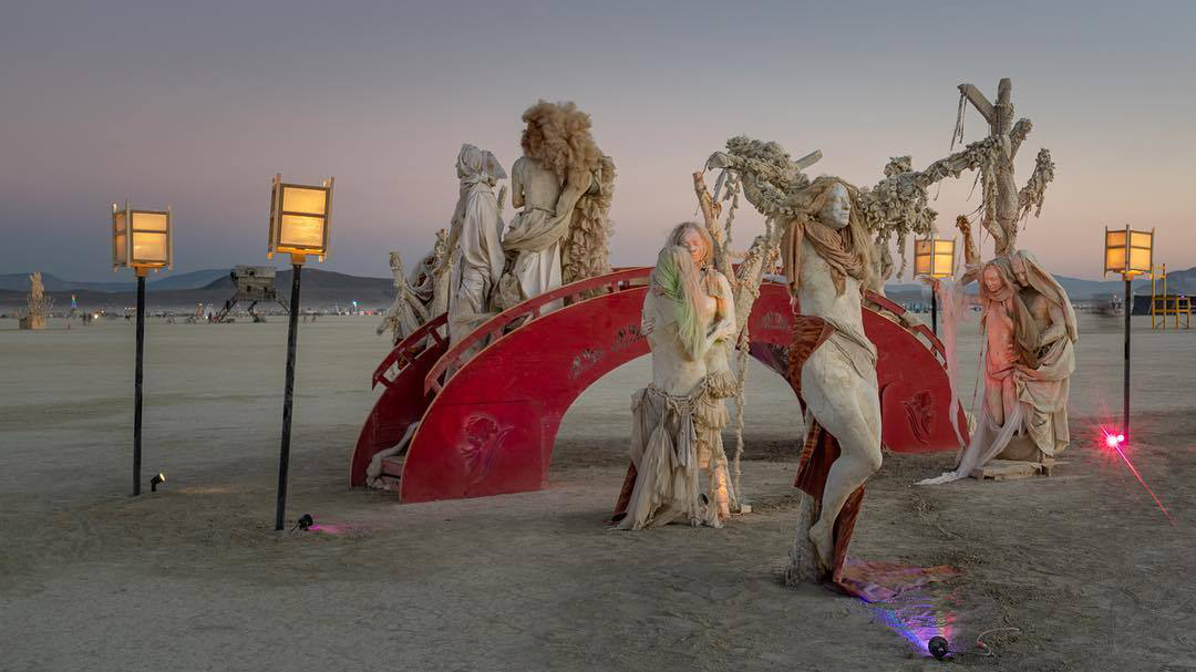 Robed figures beside a glowing red arched bridge on the playa at dusk
