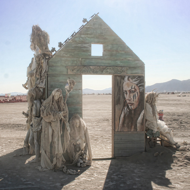 Soul Refuge installation, a weathered wooden house frame with robed figures and a painted portrait, set on the playa